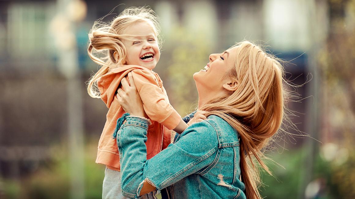 Matka trzyma w górze roześmianą pięciolatkę z kucykami