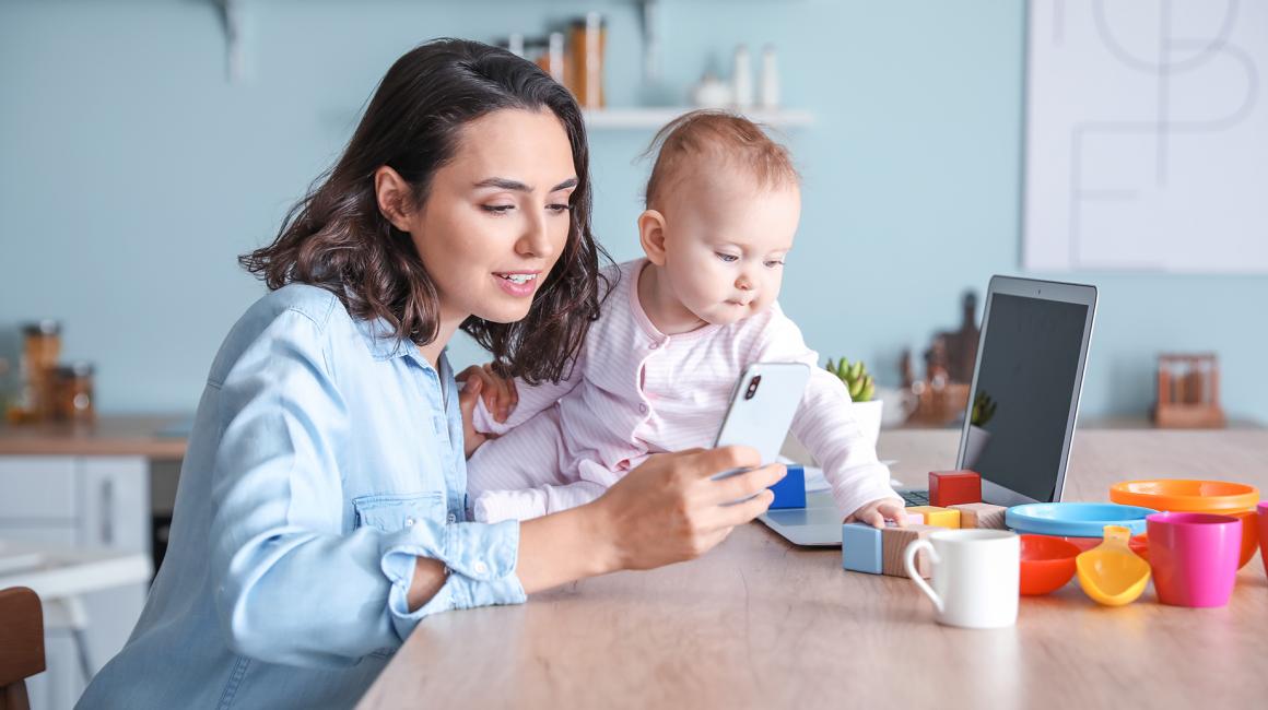 Mama z niemowlakiem, trzyma komórkę, przed nią komputer