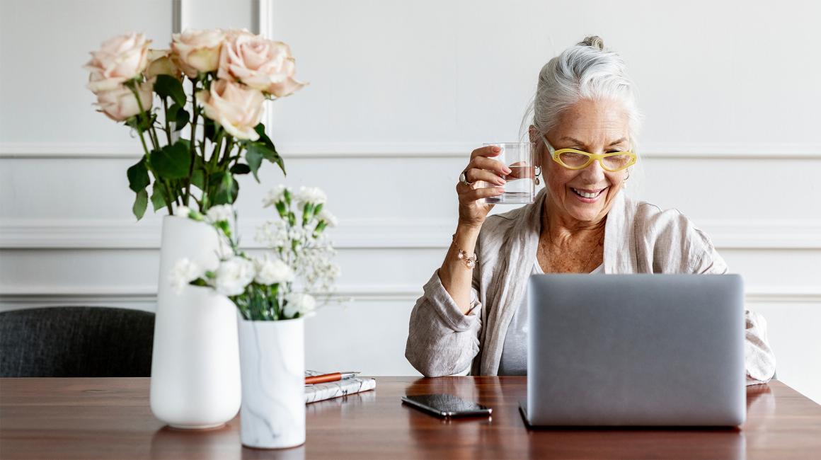 Starsza pani w koku przed laptopem, na stoliki w wazonach dwa bukiety: róże i goździki