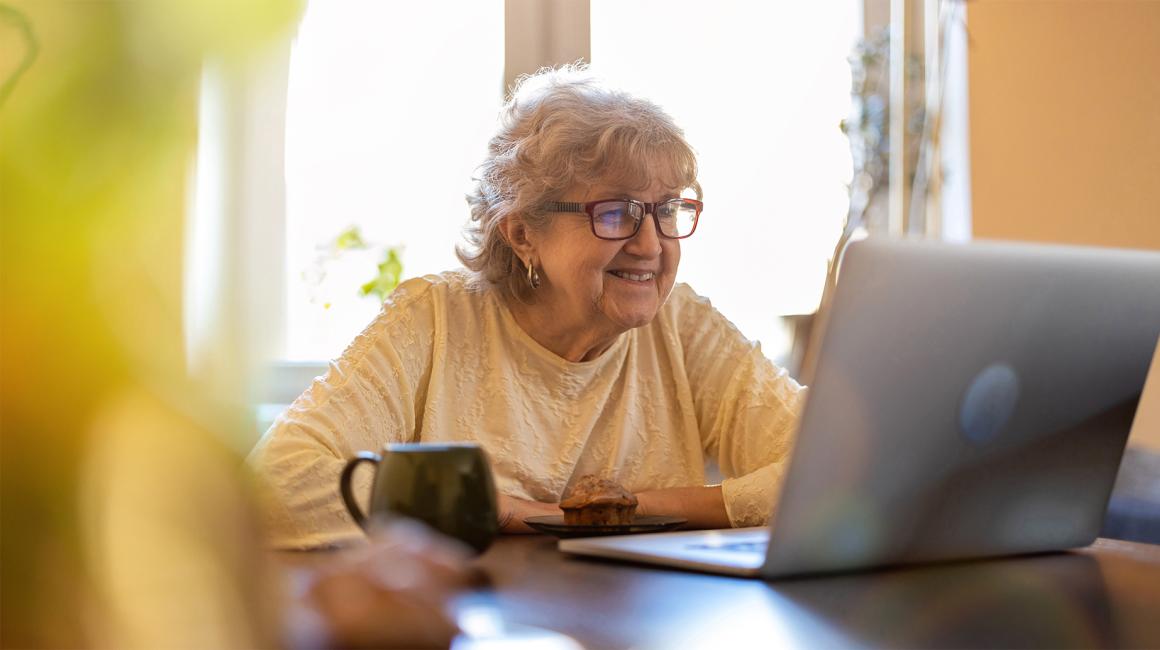 starsza pani w swetrze przy stole przed laptopem i kubkiem herbaty