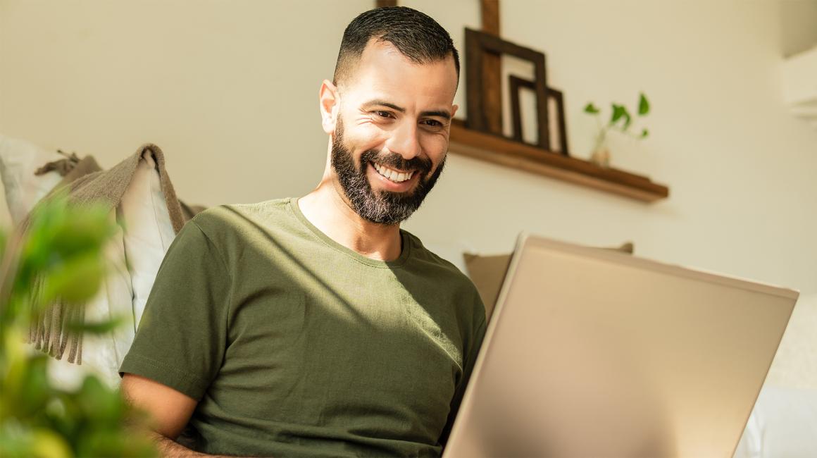 brodaty mężczyzna z laptopem siedzi, za nim widać półkę z wazonem