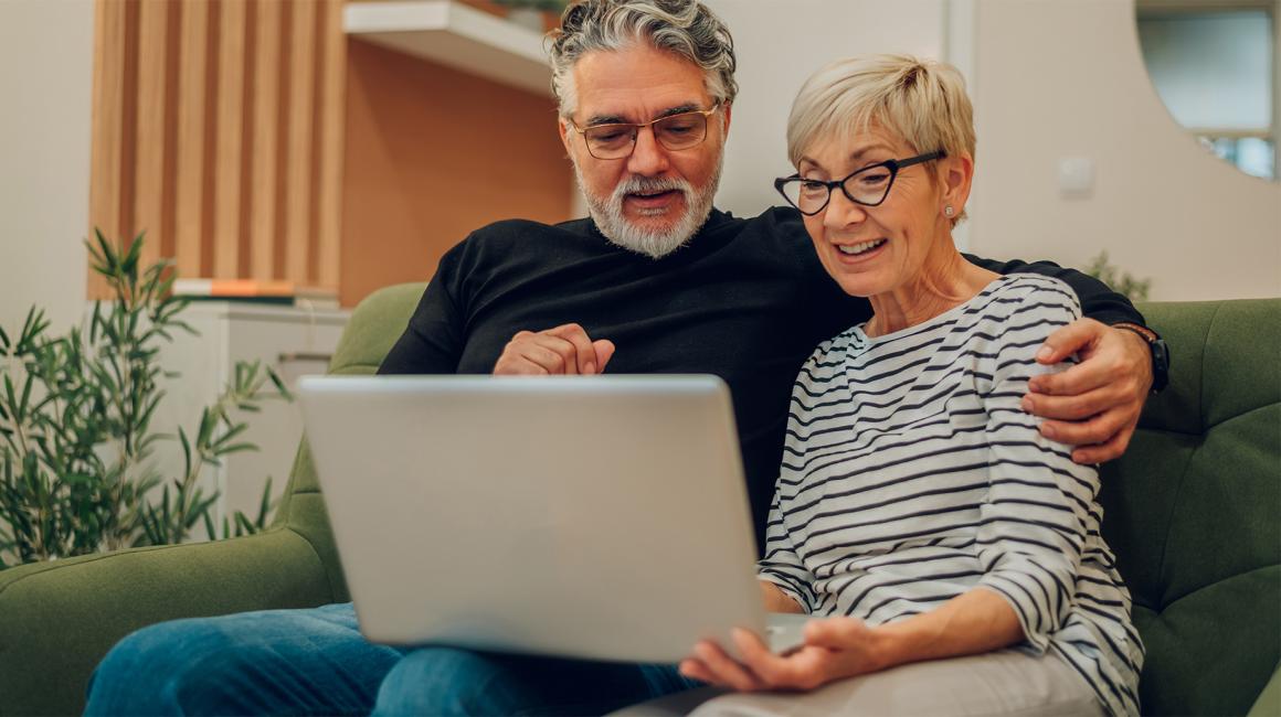 Para 50latków z laptopem na kanapie, on ją obejmuje ramieniem 