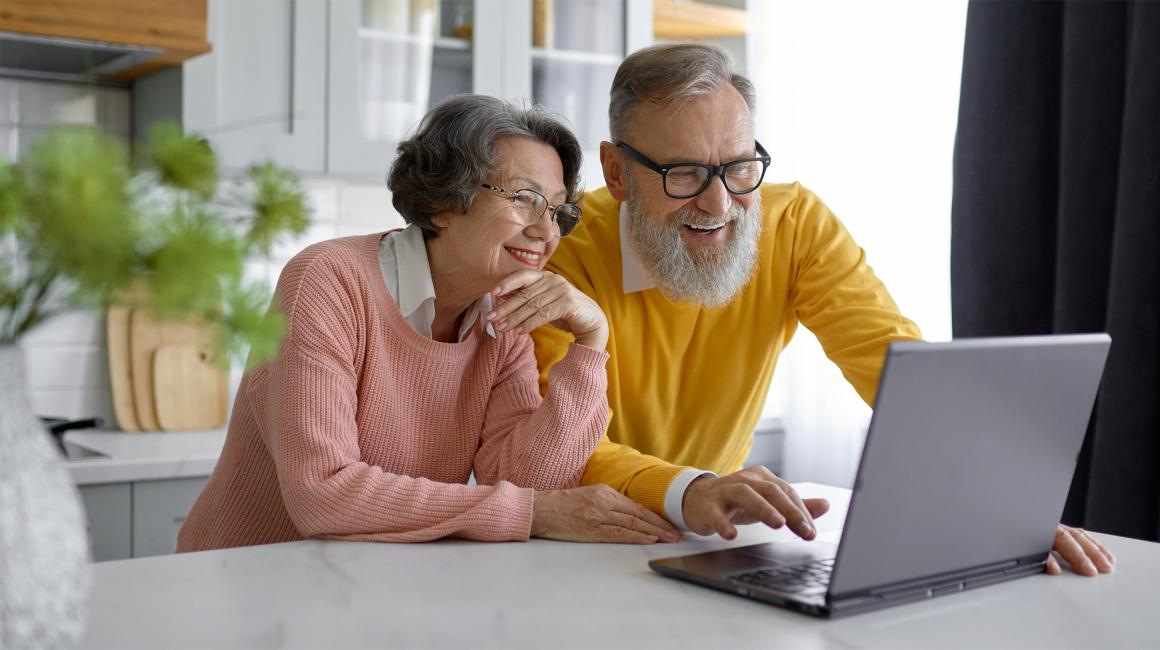 Para w starszym wieku, on z siwą broda, uśmiechnięci, w kuchni przy stole, na stole laptop
