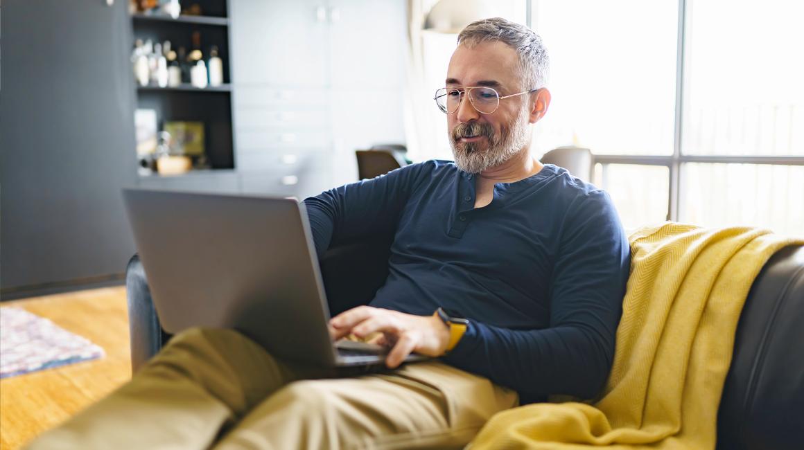 Pan z siwą brodą na kanapie z laptopem na kolanach