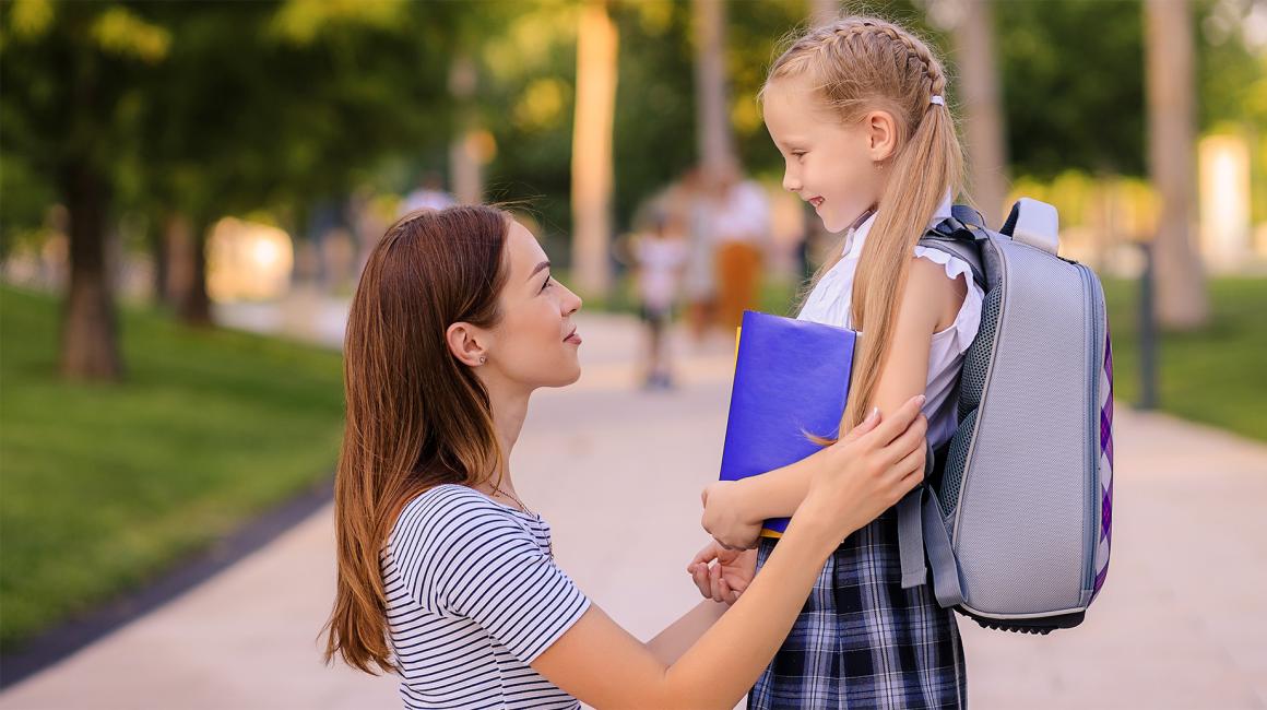 mama kuca i trzyma za ręce córkę z tornistrem, patrza na siebie z uśmiechami