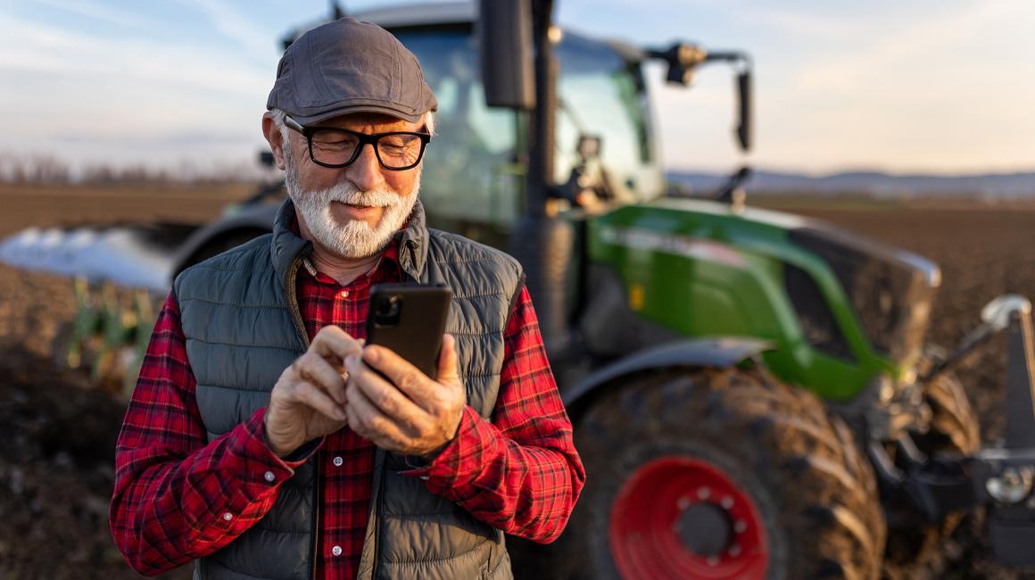 Pan z siwą brodą z komórką, w tle traktor