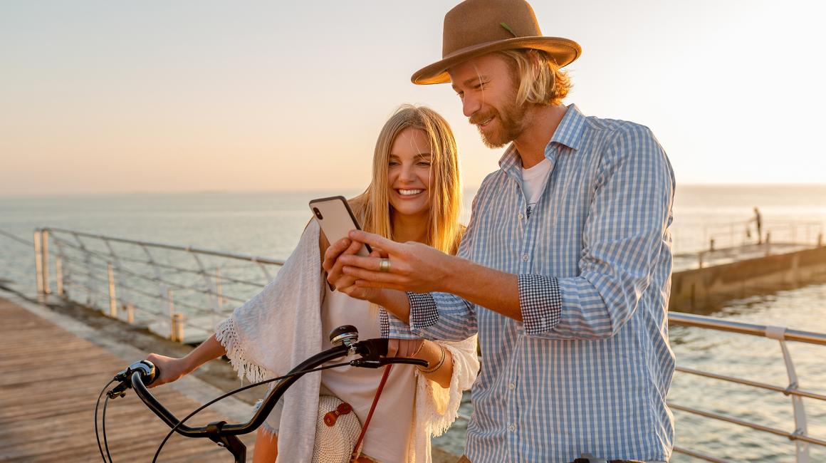 Dziewczyna i chłopak stoją na molo . Dziewczyna trzyma oburącz kierownicę roweru. Mężczyzna pokazuje jej coś na telefonie. W tle morze.