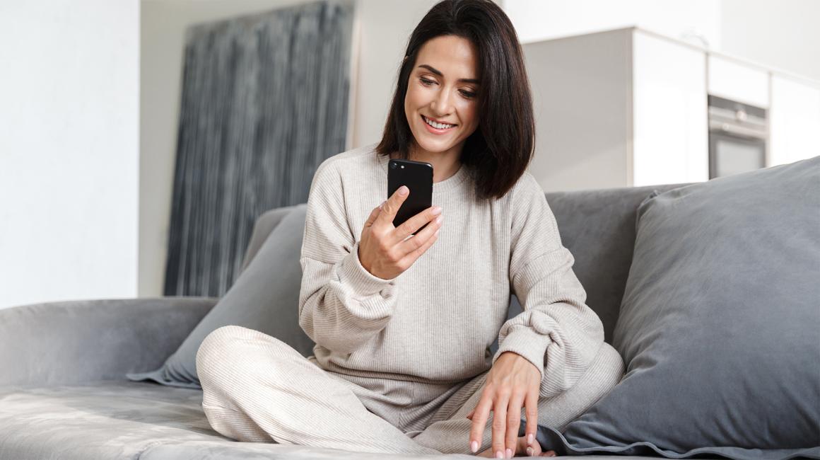 Kobieta na kanapie siedzi po turecku, ma komórkę