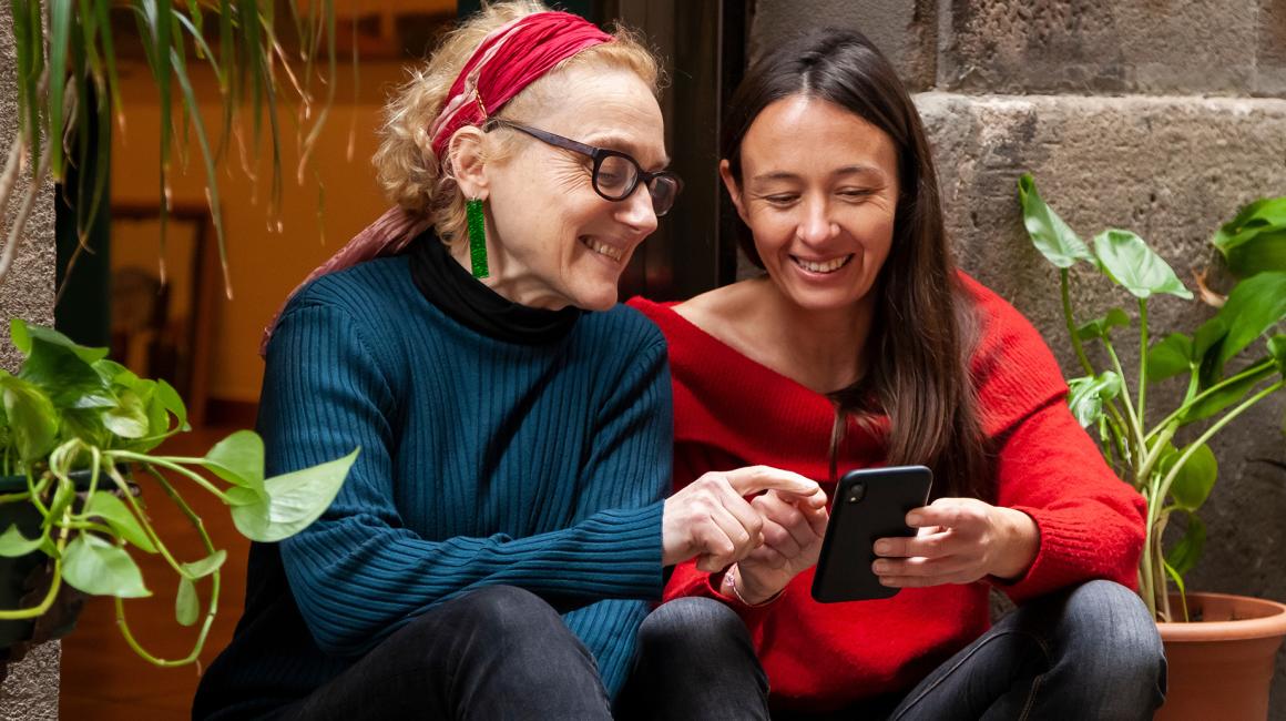 Dwie kobiety siedzą na schodach wśród zieleni i sprawdzają coś na telefonie.