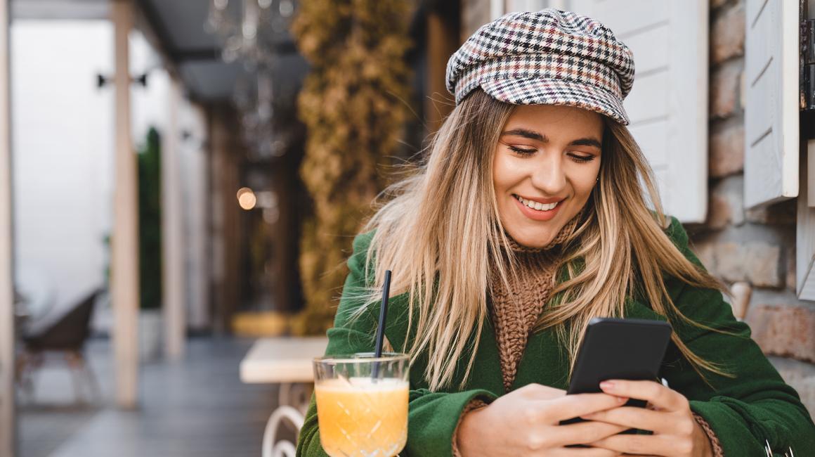 diewczyna w kyrtce i czapce z daszkiem pije kawę przy stoliku na ulicy i patrzy w komórkę