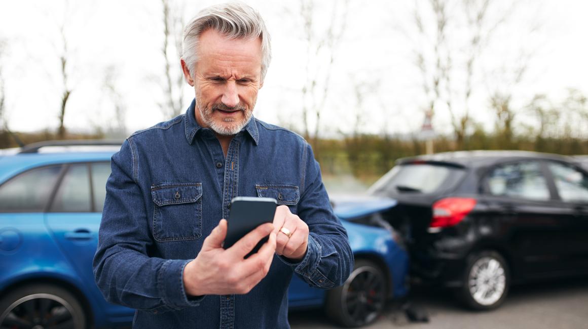 w tle zderzenie 2 aut mężczyzna z siwą bródką patrzy na telefon