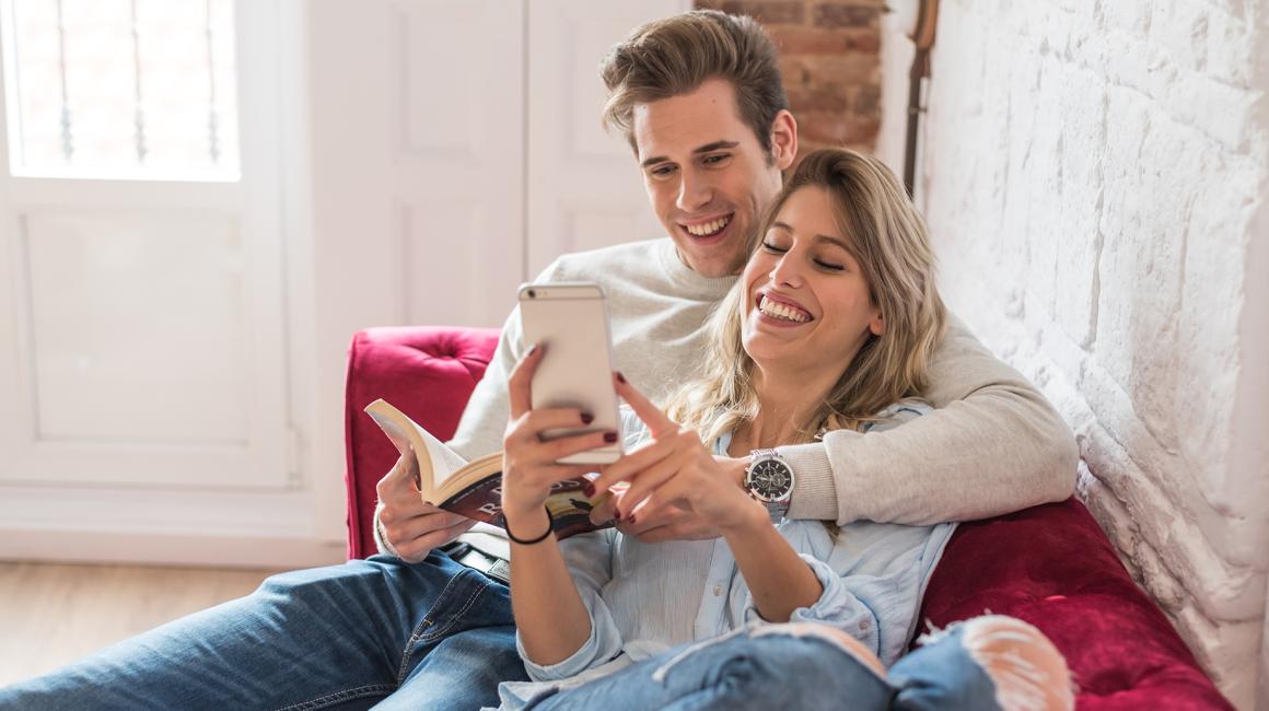 para siedzi wtulona na kanapie, on z ksiazka, ona z komórką mu coś pokazuje z uśmiechem