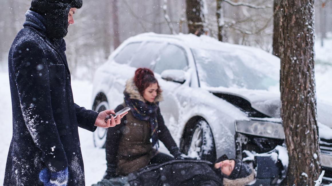 śnieg, wypadek samochodowy, jedna osoba trzyma komorke, druga udziela pomocy poszkodowanemu