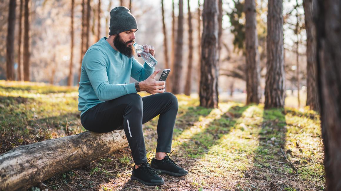 brodaty mężczyzna w lesie z butelką wody,siedzi na pniu i patrzy na komórkę