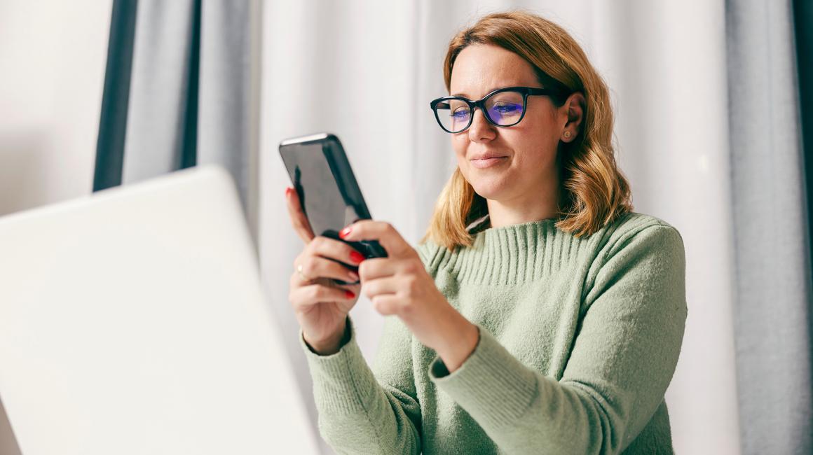 30latka z komórką, na którą patrzy, przed nią laptop