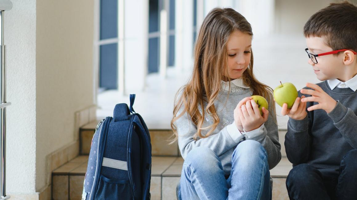 Dziewczynka i chłopiec z podstawówki siedzą na schodach w szkole, trzymają zielone jabłka i patrzą na nie.