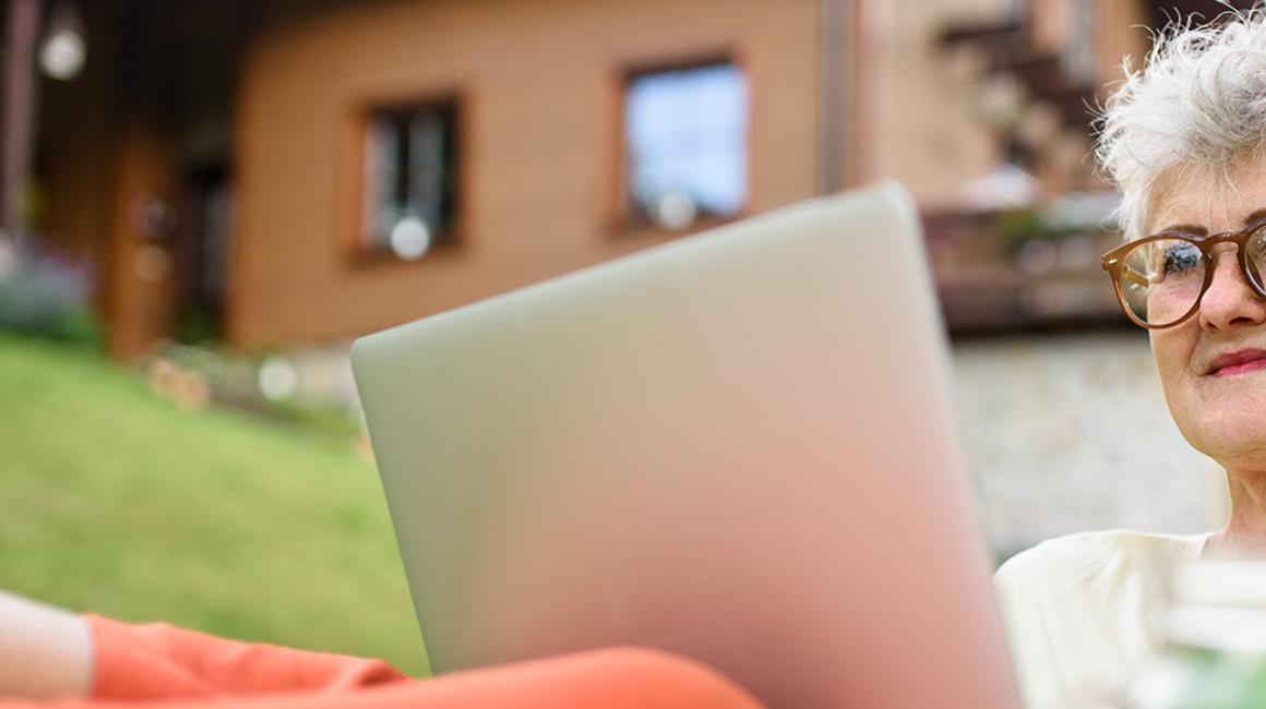 Siwa pani z laptopem przed domem w górach, laptop na kolanach, nogi na stole, na stole arbuz, w tle dom na stoku