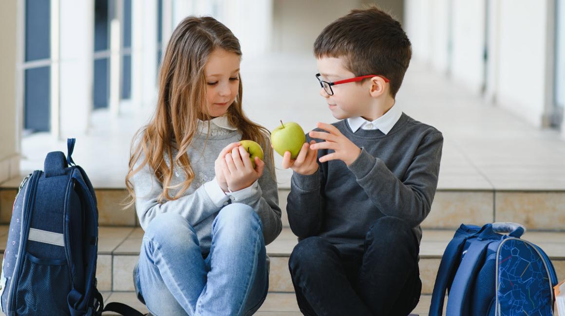 Dziewczynka i chłopiec z podstawówki siedzą na schodach w szkole, trzymają zielone jabłka i patrzą na nie.