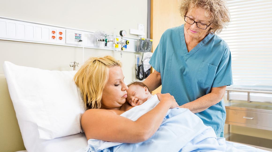 Kobieta lezy z noworodkiem, stoi położna