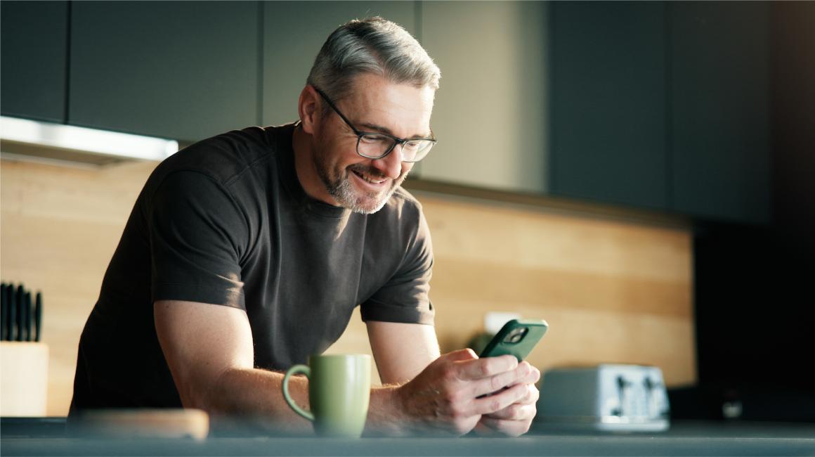 Pan z komórką w ręku, oparty łokciami o stół w kuchni