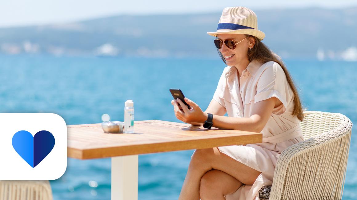 Kobieta siedzi prrzy stoliku, na stole butelka wody, ona w kapeluszu i ciemnych okularach, z komórką, w tle morze