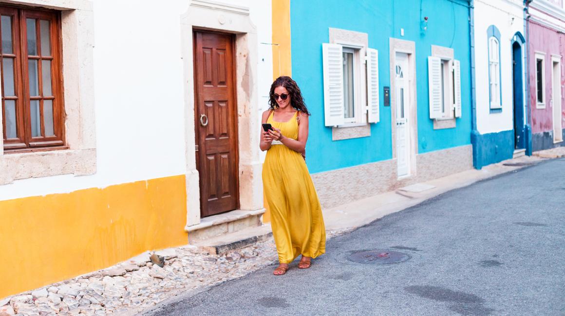 kobieta w żółtej sukience na tle biało-żółtego i bialo-niebieskiego domu, z komórką