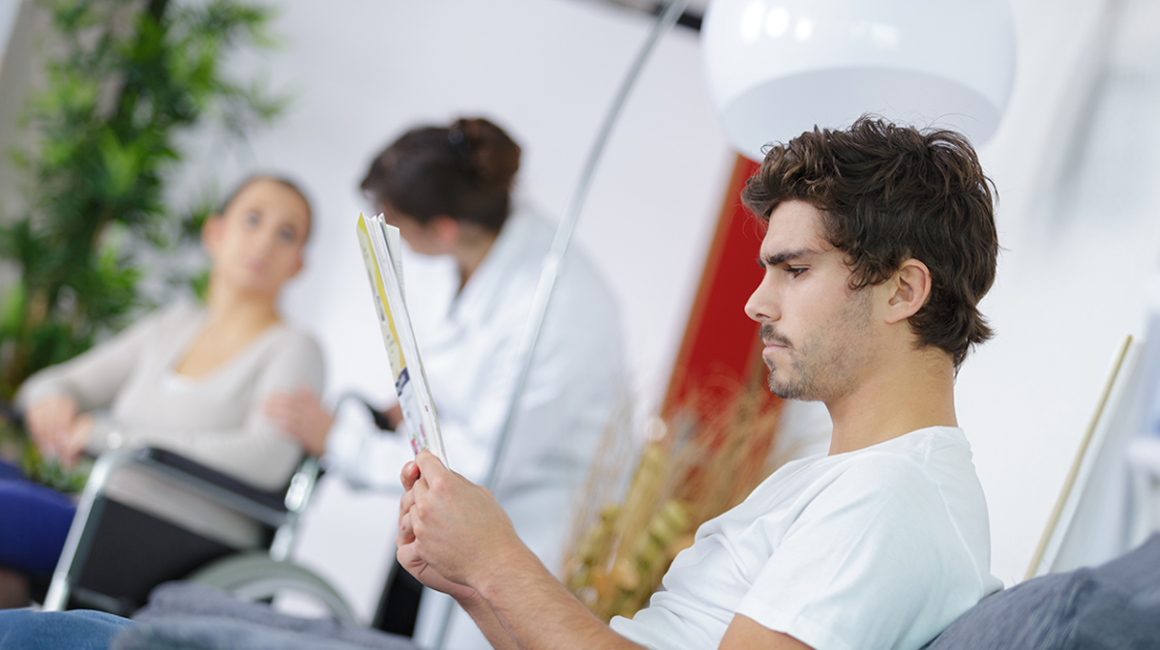 Młody mężczyzna czyta broszurę informacyjną. W tle osoba na wózku inwalidzkim i lekarka.