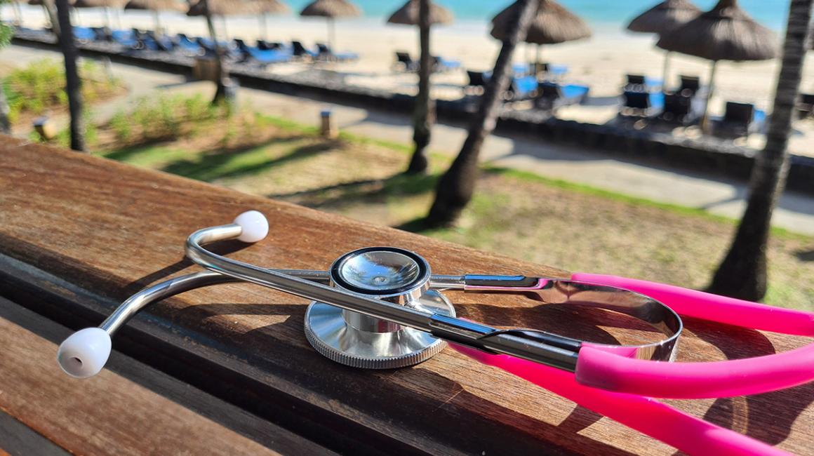 Różowy stetoskop leży na ławce w słońcu, w tle palmy i parasole na plaży