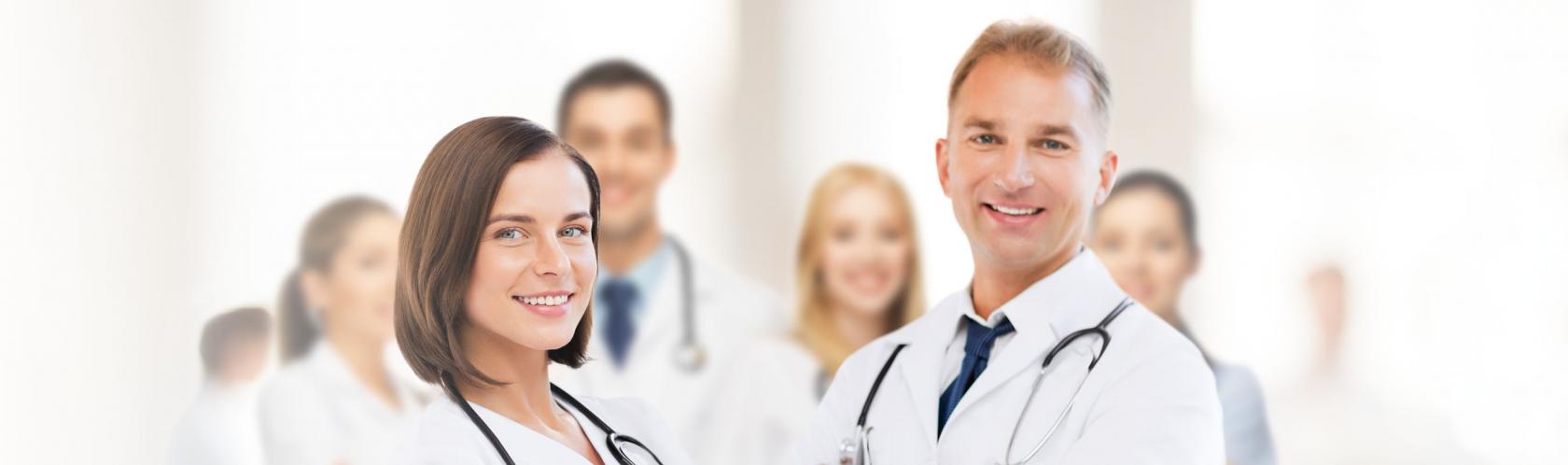 Na zdjęciu jest grupa usmiechniętych lekarzy w kitlach. Po lewej stronie stoi kobieta, po prawej mężczyzna, reszta niewyraźna w tle.