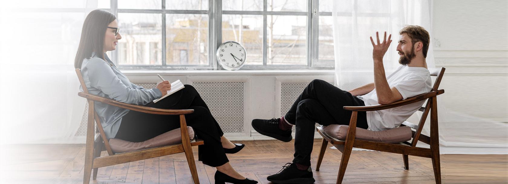 Mężczyna i kobieta siedzący naprzeciwko siebie. Kobieta ma notes i długopis. Mężczyzna opowiada z gestykulacją 