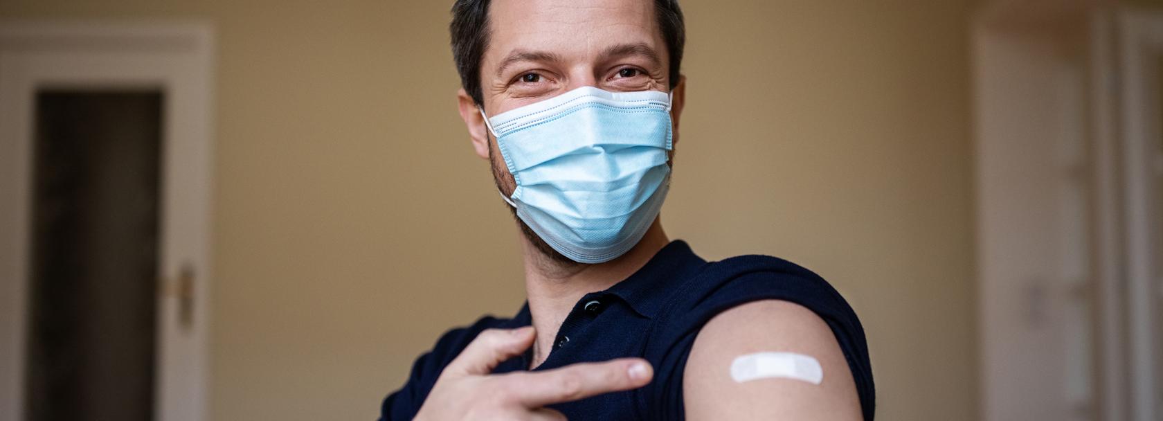 młody mężczyzna w maseczce pokazuje na ramię z plastrem po szczepieniu