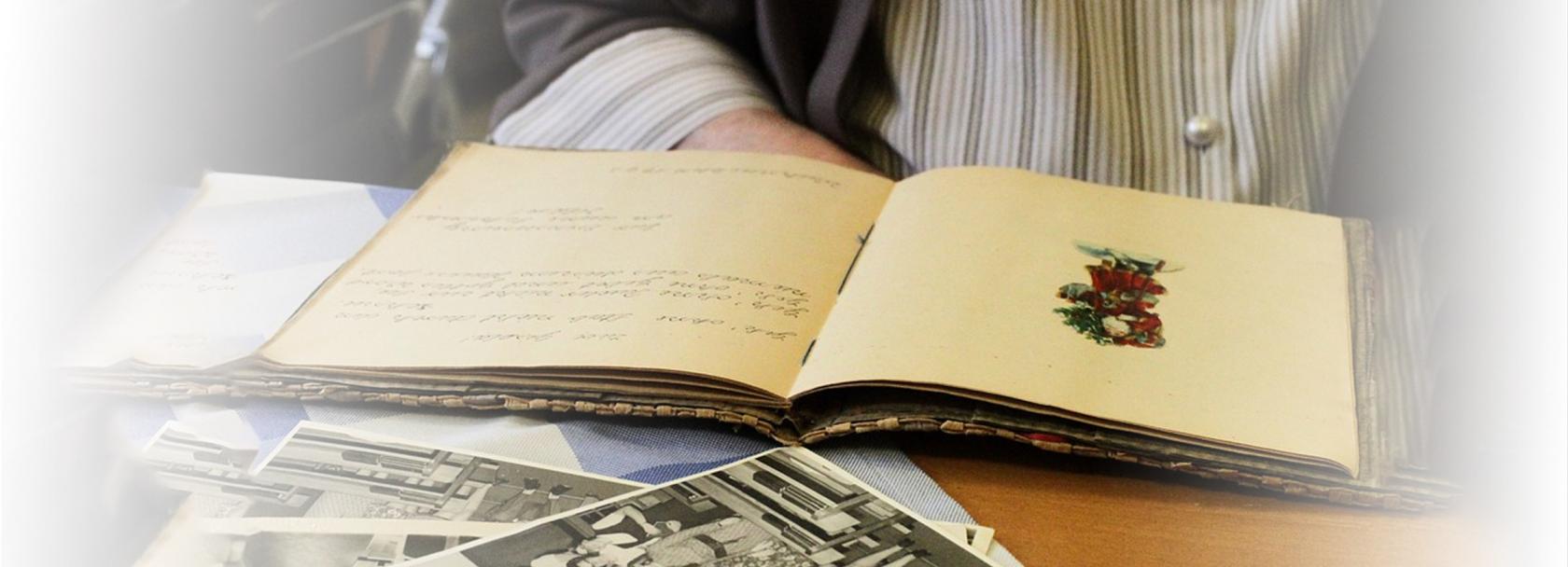 Leżące na stole lub biurku stare zdjęcia i otwarty pamiętnik. W tle zarys siedzącej osoby