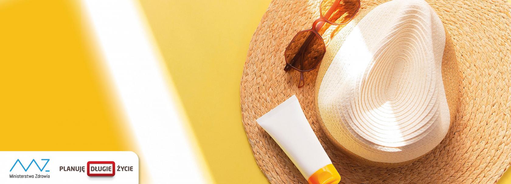 na żółtym słonecznym tle słomkowy kapelusz, na nim okulary i krem do opalania