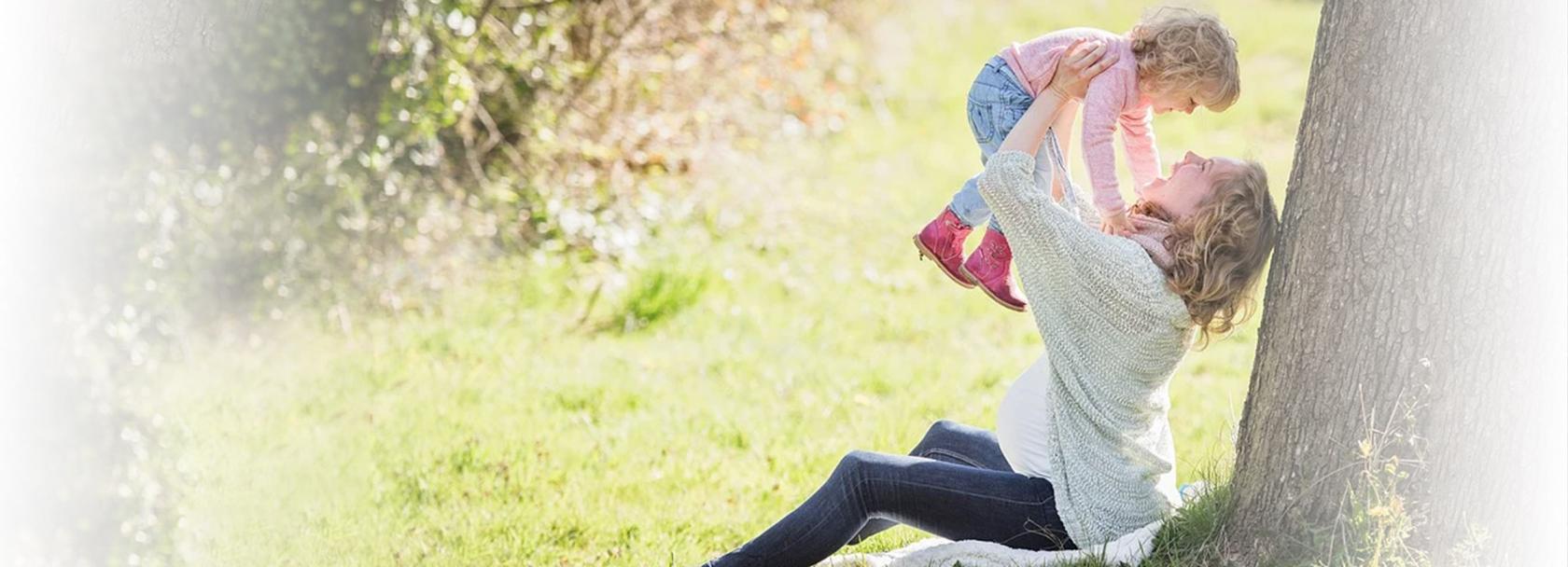 Kobieta w ciąży siedząca przy drzewie i trzymająca w górze dziecko