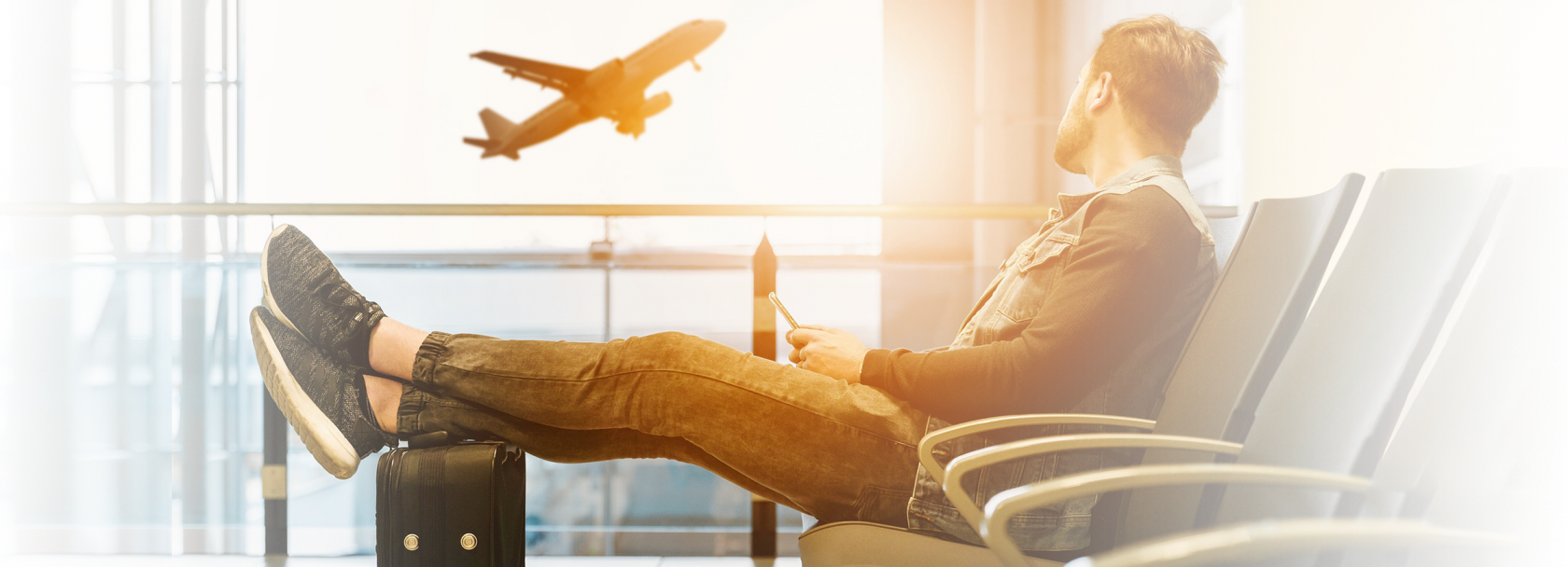 Mężczyzna siedzący w poczekalni lotniska. Uniesione nogi opiera na walice. Patrzy na widoczny za oknem startujący samolot