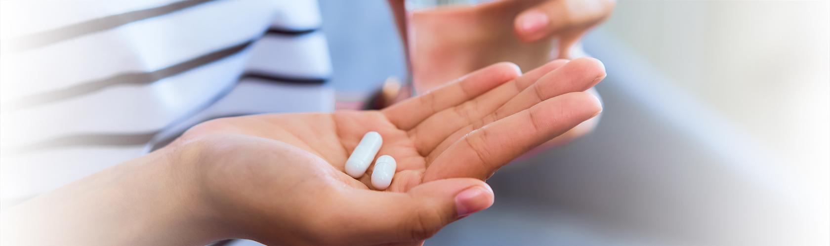 Wykadrowane ręce siedzącej osoby. W prawej ręce 2 tabletki, a w lewej szklanka z wodą