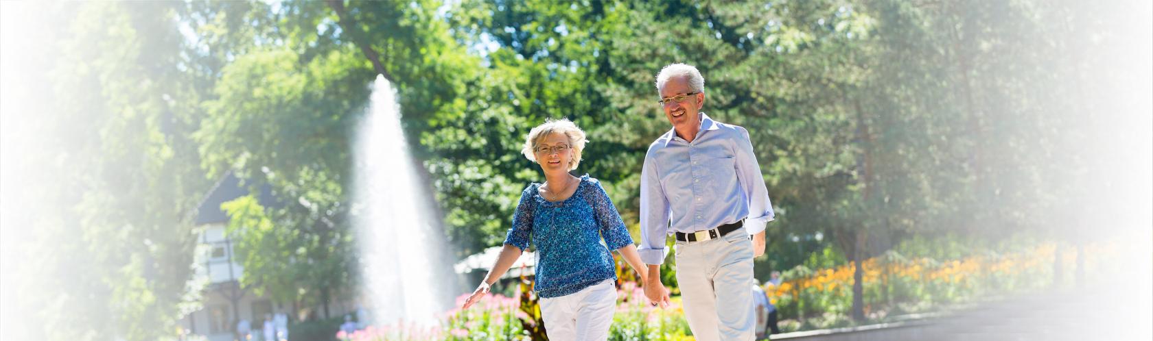 Para seniorów spacaruje po parku, w tle fontanna 