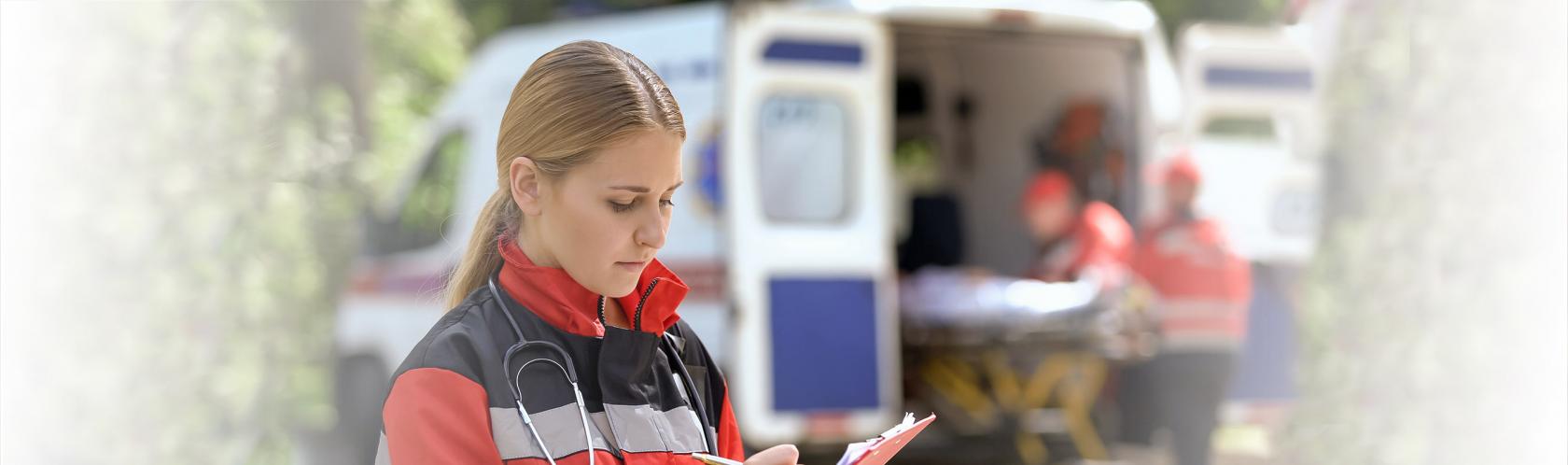 Na pierwszy planie kobieta zapisująca informacje w notesie. Na drugim planie karetka, z której 2 osoby wyjmują nosze.