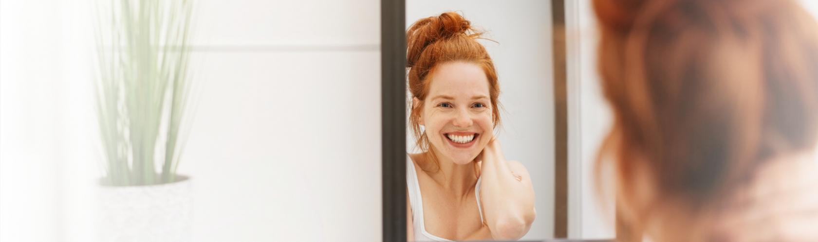 Uśmiechnięta kobieta stoi w łazience i przegląda się w lustrze.