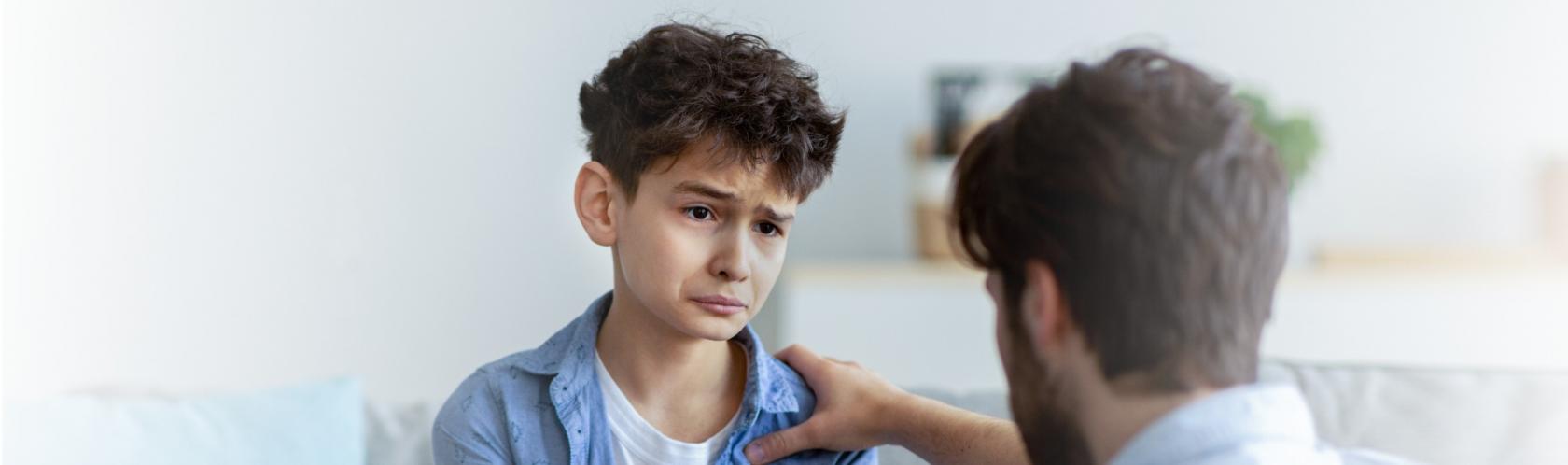 Trudna rozmowa ojca z synem. Chłopiec jest zasmucony, ojciec go wspiera, trzymając rękę na ramieniu.