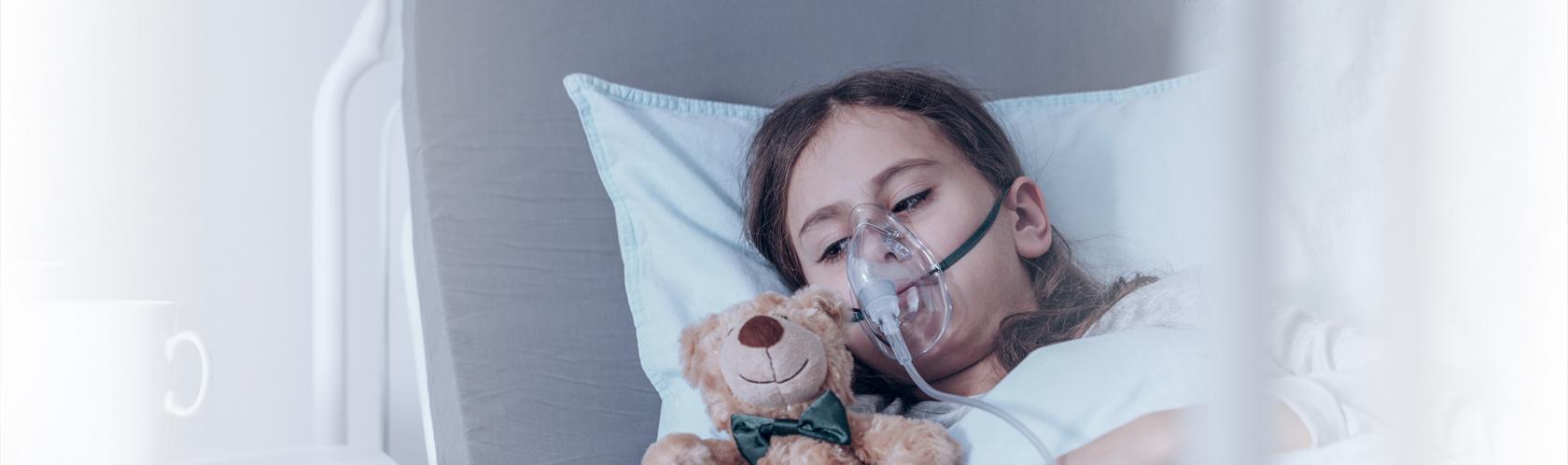 Dziecko w masce tlenowej na szpitalnym łóżku. Przytula misia.