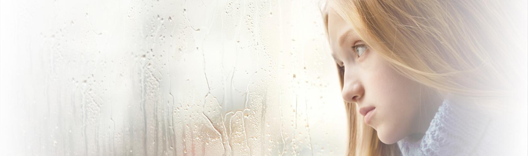 Smutna dziewcynka z długimi blond włosami patrzy przez szybę w dal. Za oknem pada deszcz.