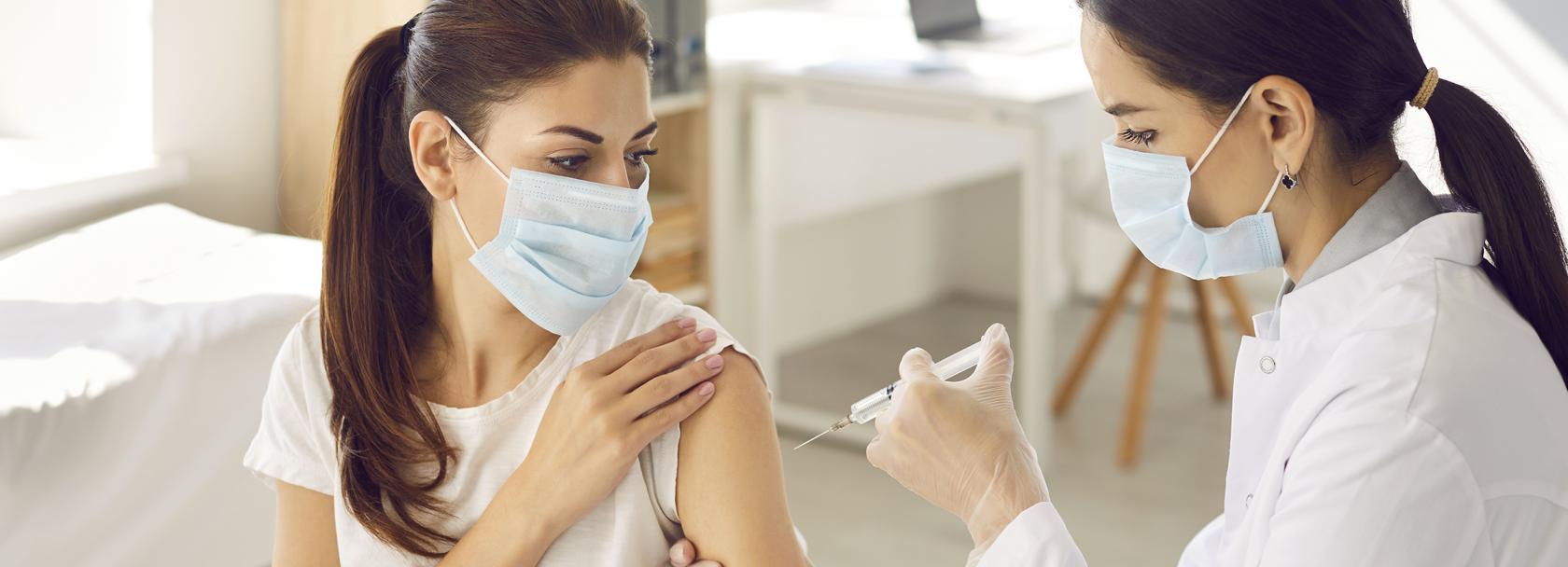 Pielęgniarka wykonuje kobiecie zastrzyk, obie w maseczkach