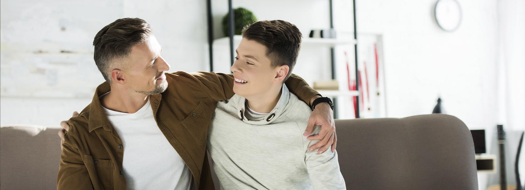 Ojciec obejmuje ramieniem nastoletniego syna, siedzą na kanapie