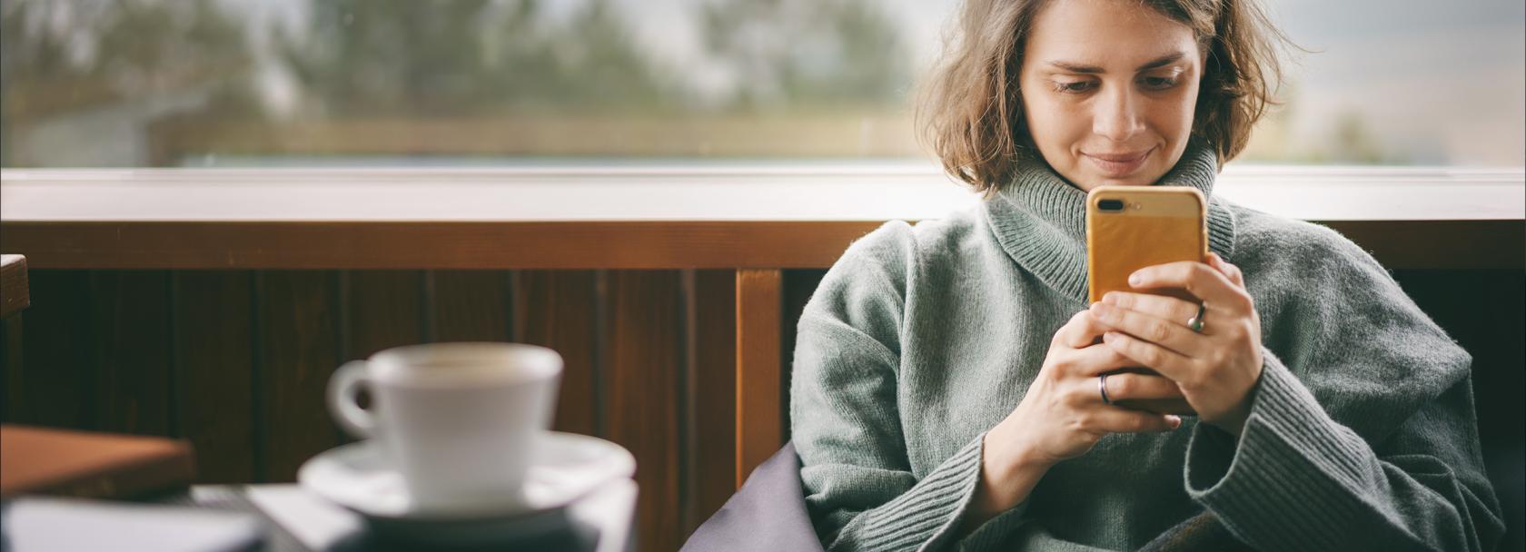 Kobieta w dresie patrzy na komórkę, przed nią kawa