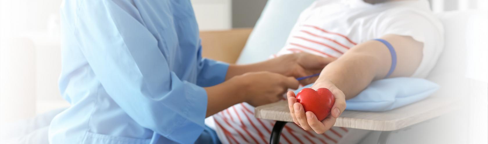 Pielęgniarka przygotowuje mężczyznę do oddania krwi. Zakłada mu opaskę uciskową. Mężczyzna trzyma w dłoni gumowe czerwone serce.