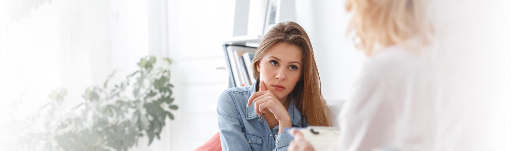 Na pierwszym planie mamy zadbaną młodą kobietę. Słucha, co mówi druga, która siedzi do nas tyłem.