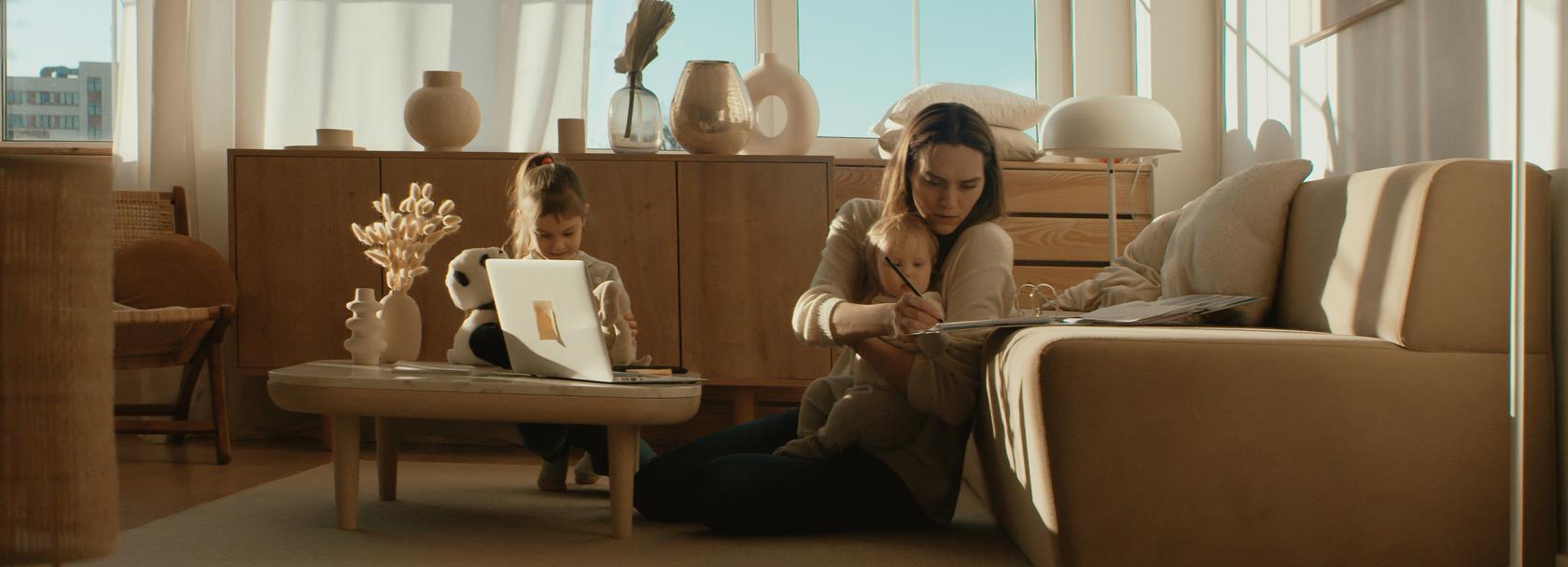 Kobieta siedzi na podłodze przy kanapie z małym dzieckiem, starsze dziecko bawi się na stoliku