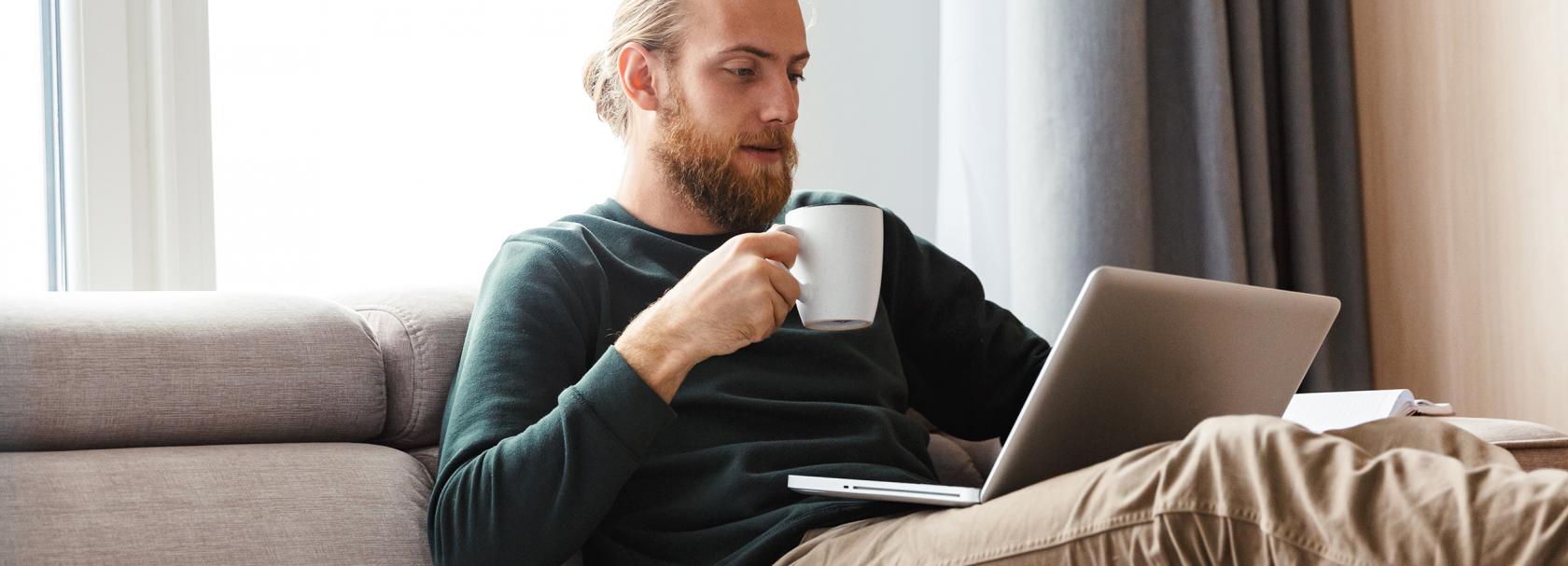 Brodaty młody facet z kubkiem na kanapie i laptopem na kolanach