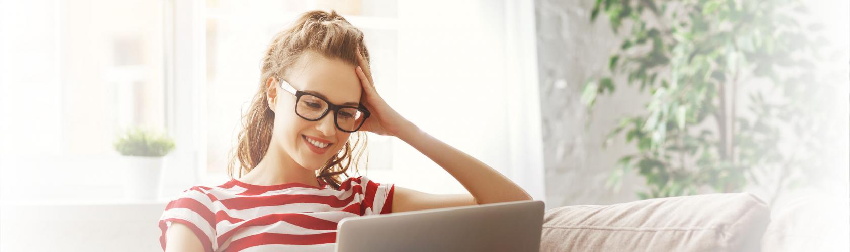 Młoda dziewczyna w okularach i koszulce w czerwone paski siedzi z laptopem na kolanach.